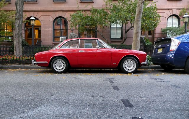 A classic red car parked on a city street with trees and buildings in the background.